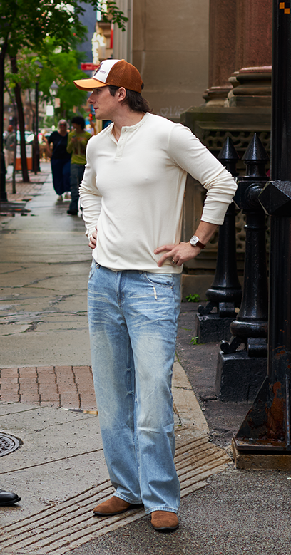Two men standing on a city sidewalk in an urban street setting. One man wears a white graphic T-shirt, dark jeans, and black shoes, while the other wears a cream long-sleeve shirt, light blue jeans, and an orange baseball cap.