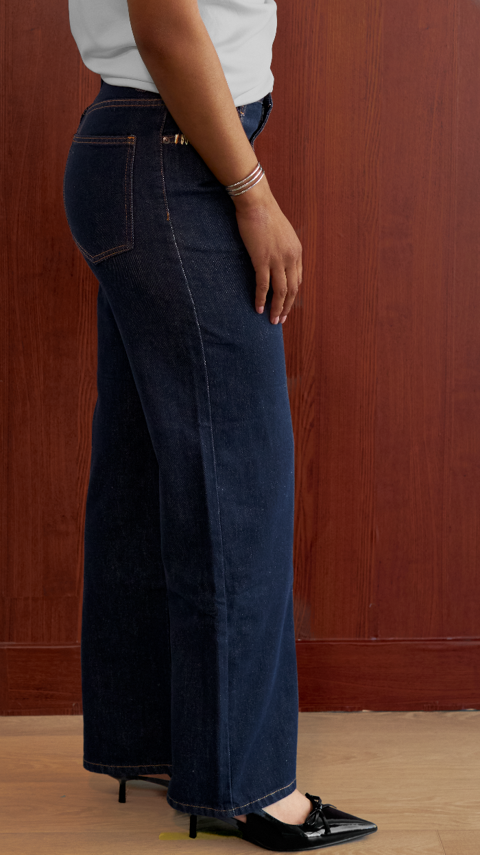 A person shown from the waist down wearing dark blue wide-leg jeans, a white top, and black pointed flats, standing against a brown wooden wall.